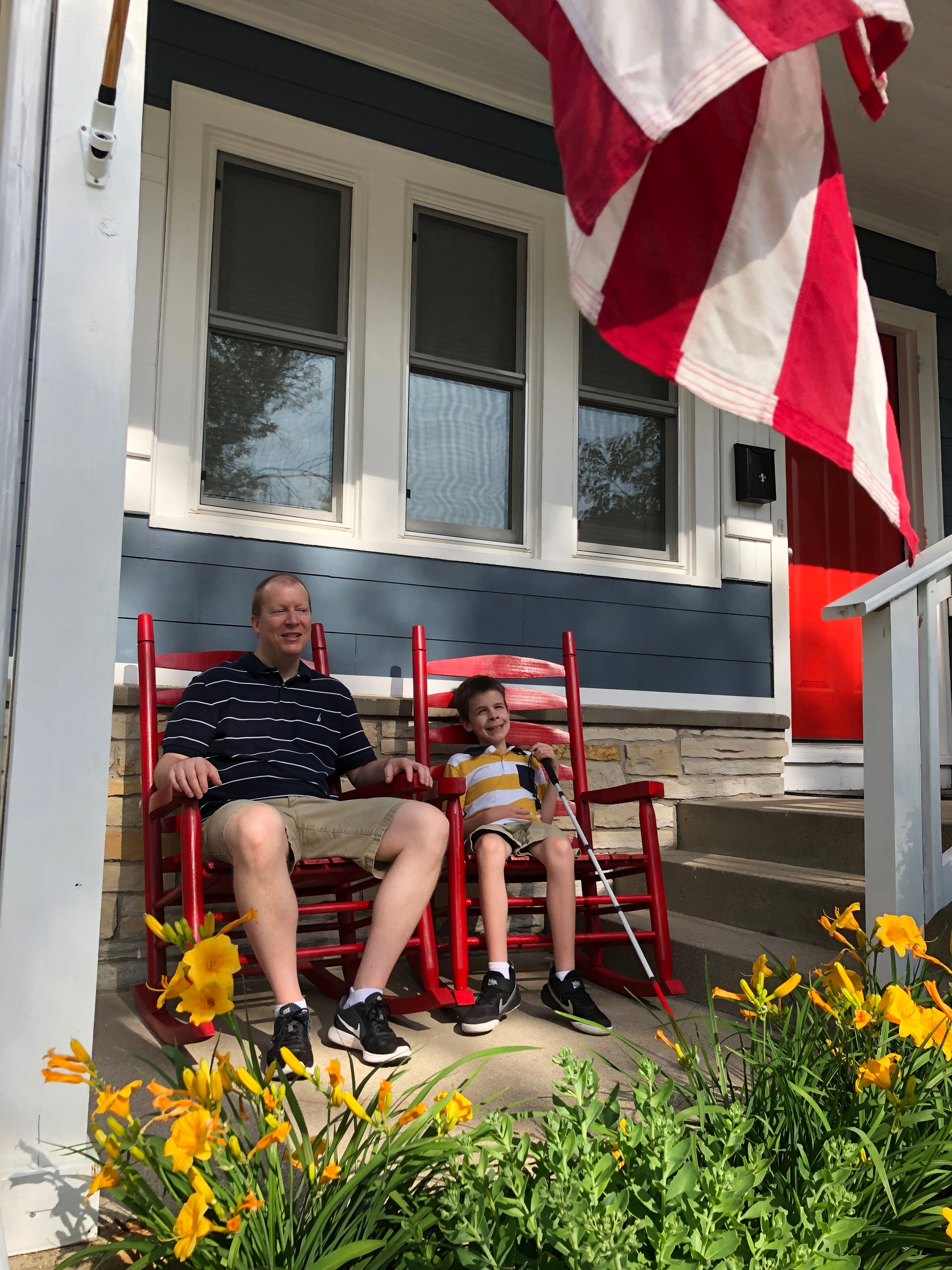 A visually impaired man and boy sit smiling in red rocking chairs on the front porch of a blue house. Flowers and an American flag are visible in the foreground.